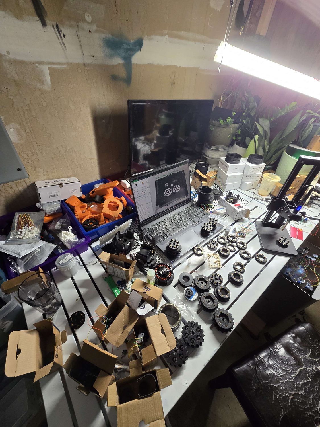 Garage workbench with a 3D printer, staged actuator parts, and a CAD actuator shaft profile open on a laptop.