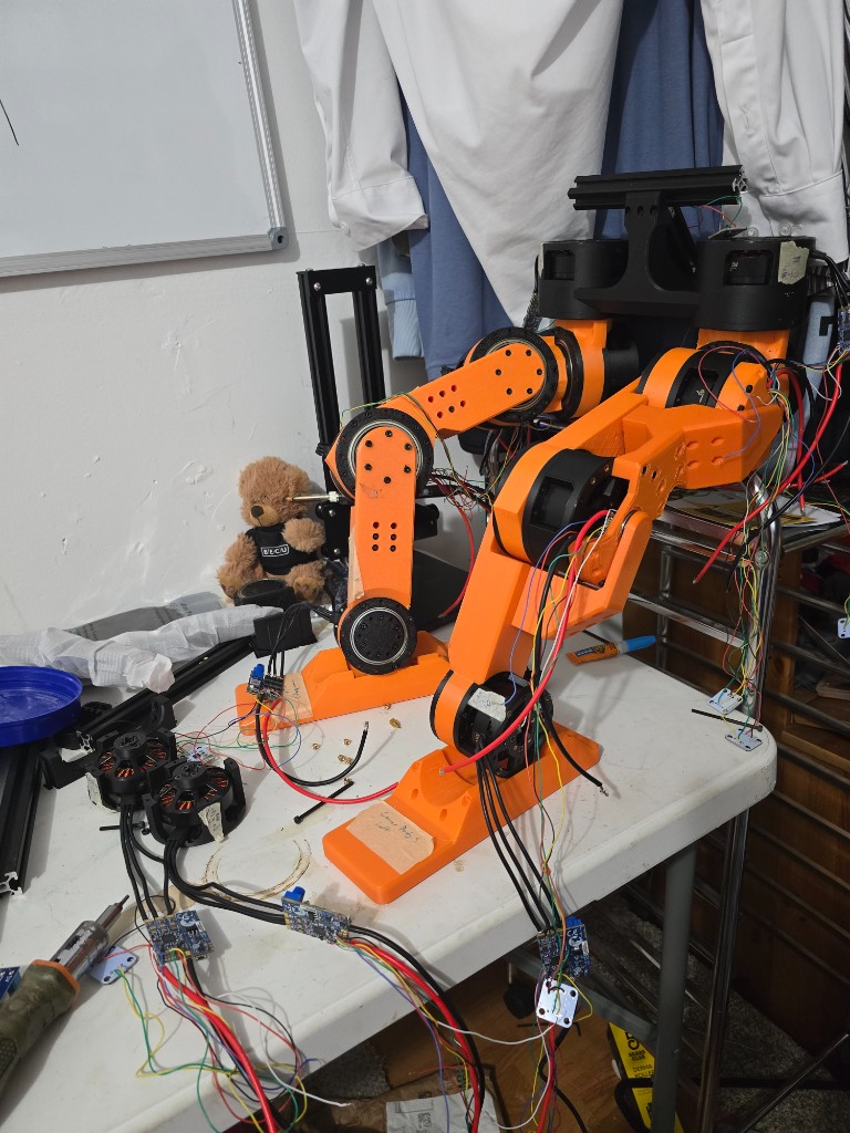 A pair of orange 3D-printed humanoid legs on a folding table, wiring still external.