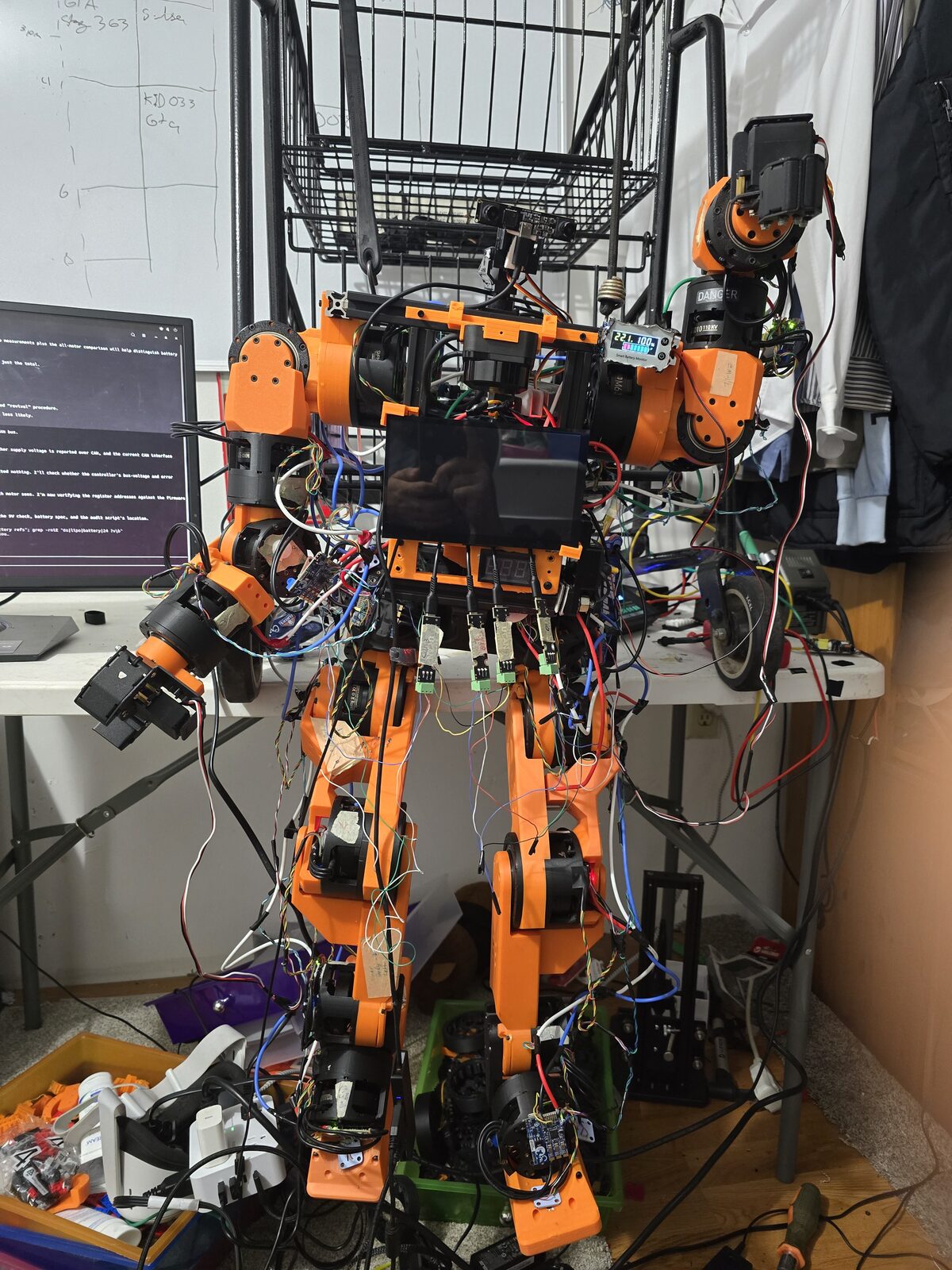 Fully assembled Berkeley Humanoid Lite hanging from a rack, chest screen on, wiring still external.